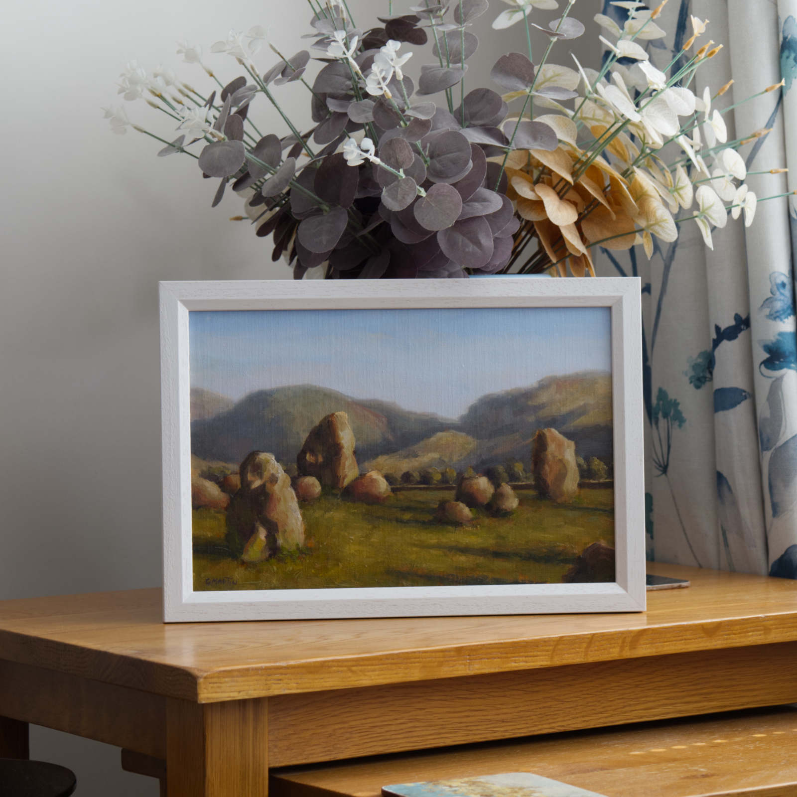 Framed landscape painting of Keswick Stone circle on a table in front of artificial flowers